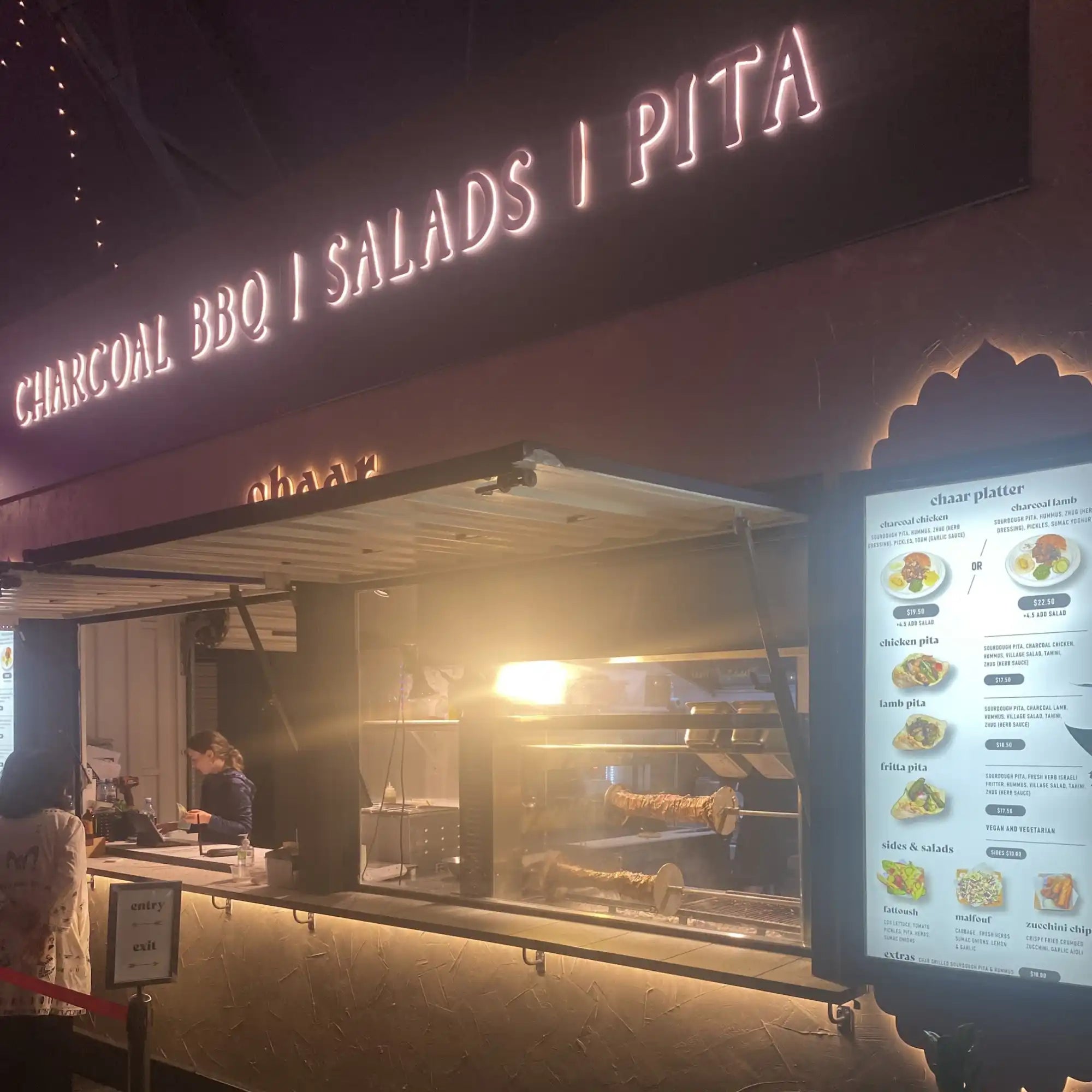 Food service counter with illuminated signage advertising charcoal BBQ, salads and pita.