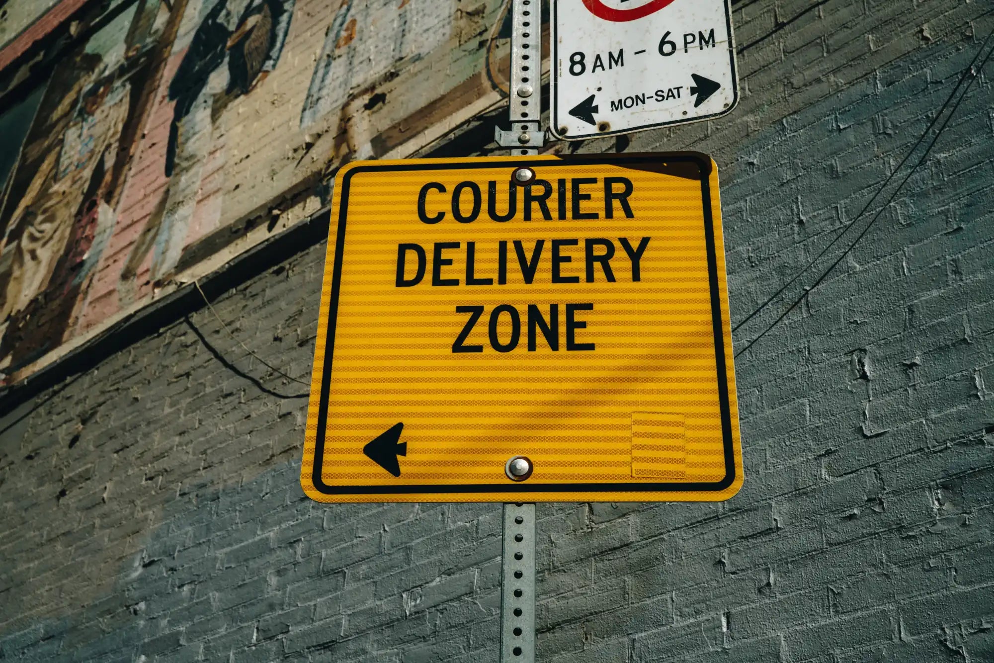 Yellow rectangular sign with black text reading ’COURIER DELIVERY ZONE’ and a directional arrow.